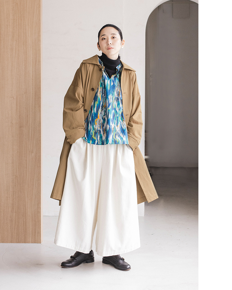 トレンチコートと青の柄ブラウス、白のワイドパンツを重ね着した人物が、木製パネルの前に立つコーディネート写真