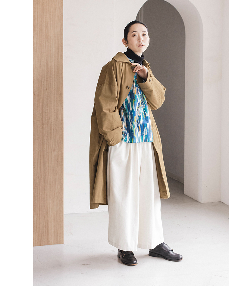 トレンチコートと青の抽象柄シャツを重ねた人物が、アーチ状のドアがある室内に立つコーディネート写真