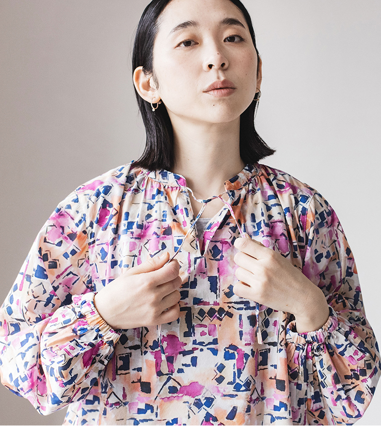 ピンク・ブルー・オレンジの抽象柄ブラウスを着た人物が、首元の紐を両手で持っているコーディネート写真
