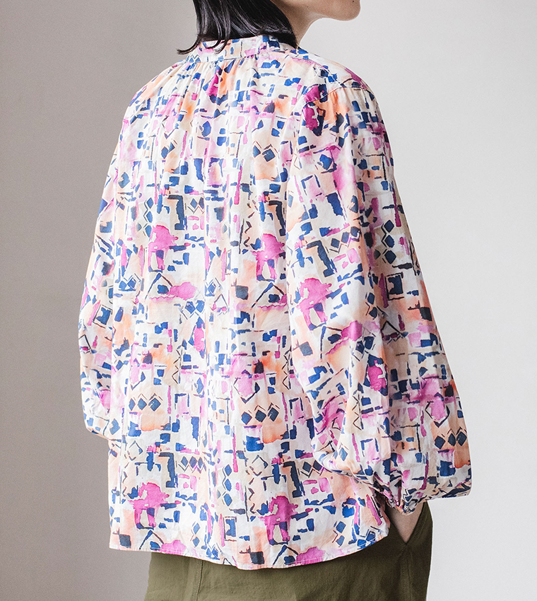 ピンク・オレンジ・ブルーの抽象柄ブラウスを着た人物の後ろ姿。ギャザー入りの襟元とボリューム袖が特徴的で、カーキのパンツを合わせたコーディネート写真