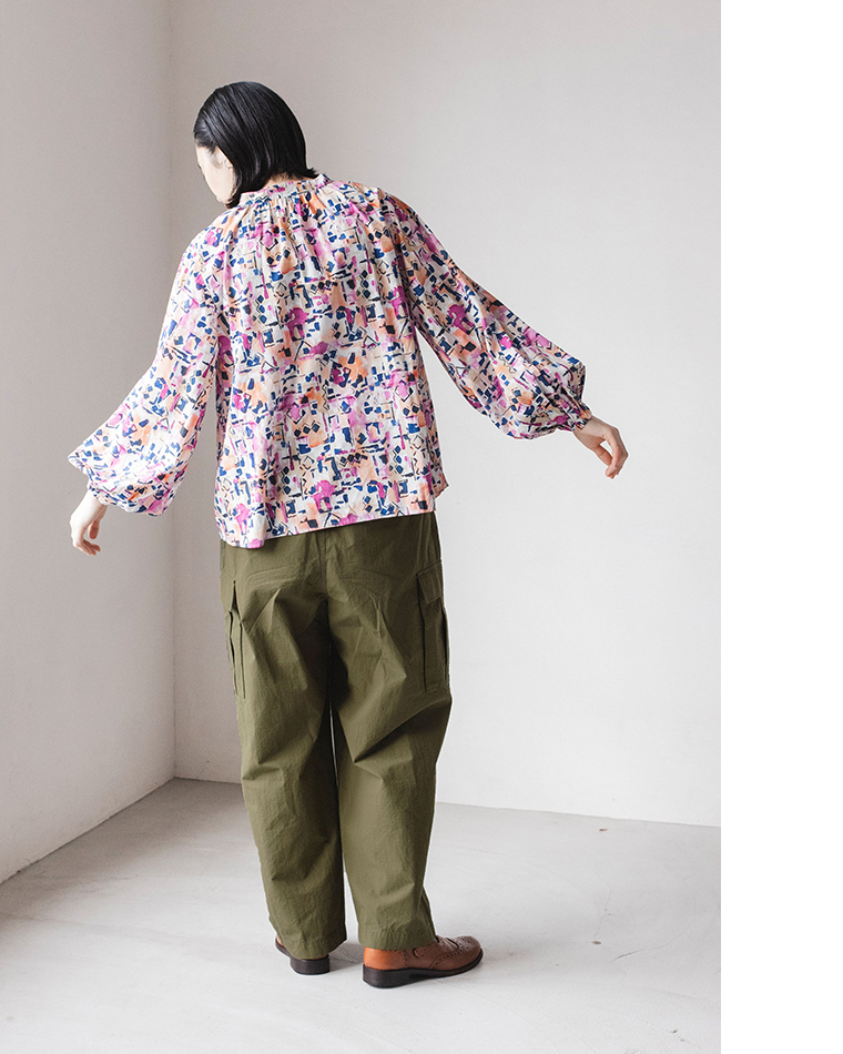 ピンク・パープル・ブルー系の抽象柄ブラウスとカーキのカーゴパンツを着た人物の後ろ姿。白い室内で立っているコーディネート写真