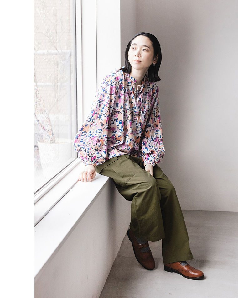ピンク・ブルー系の抽象柄ブラウスとカーキのワイドパンツを着た人物が、窓辺に腰かけて外を見つめるコーディネート写真