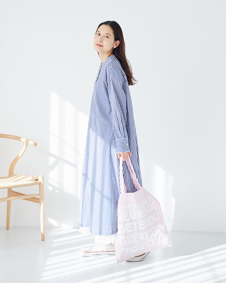 ブルーとホワイトのストライプ柄ワンピースを着用し、ピンクのトートバッグを持った後ろ姿のコーディネート（白スニーカー・室内・自然光）