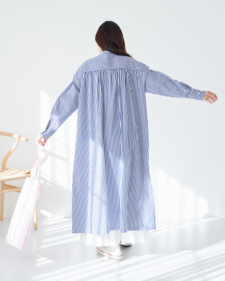 ブルーとホワイトのストライプ柄ワンピースを着用し、ピンクのトートバッグを持った後ろ姿のコーディネート（白スニーカー・白いレイヤード・明るい室内）