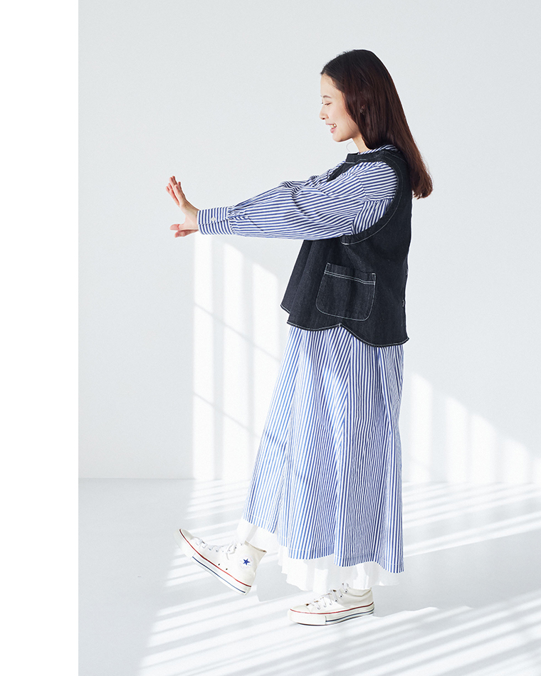 ストライプ柄ワンピースにデニムベストを重ね、白のハイカットスニーカーを合わせた横向きのコーディネート（明るい室内・ブラインドの影）