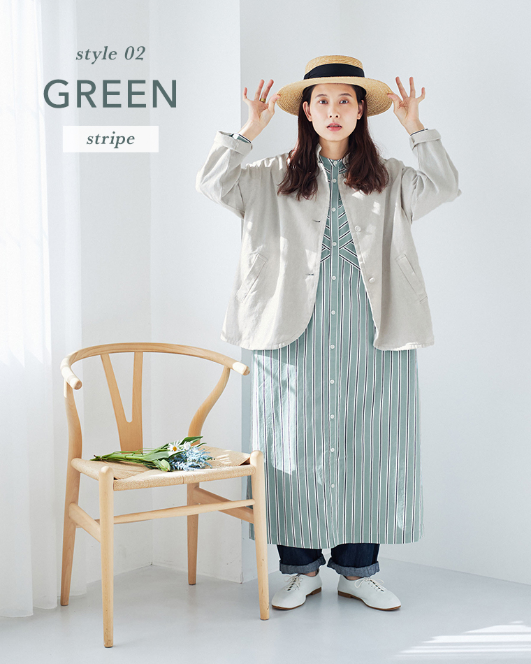  グリーンとホワイトのストライプワンピースにベージュのジャケットとストローハットを合わせた春夏コーディネート（室内・白い壁・花を置いた椅子あり）