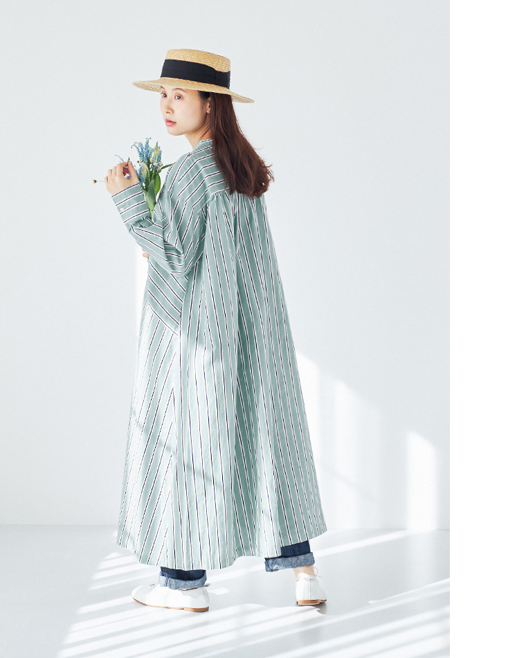 グリーン・ホワイト・ブラックのストライプワンピースにデニムパンツを重ね、ストローハットと白いシューズを合わせたコーディネート（青い花のブーケを手にした室内スタイル）