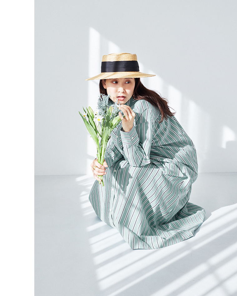 グリーンとホワイトのストライプワンピースを着用し、ストローハットをかぶって花束を持つ人物がしゃがんだポーズ（明るい室内・ブラインドの影）