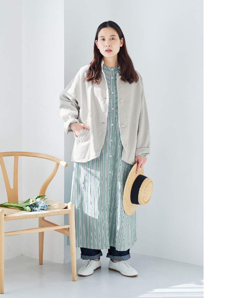 グリーンとホワイトのストライプワンピースにベージュのジャケットとデニムパンツを重ねた春のコーディネート（白いレースアップシューズ・ストローハットを手に持つ・花を置いた椅子）