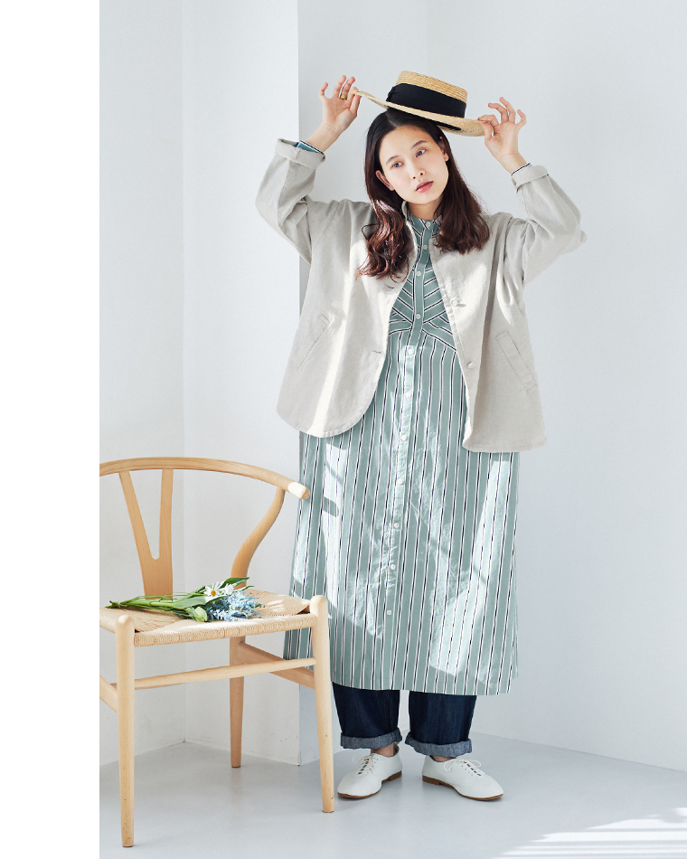 グリーンとホワイトのストライプワンピースにベージュのジャケットとデニムパンツを重ねたコーディネート（白い靴・ストローハットを頭上に持ち上げるポーズ・花を置いた椅子）