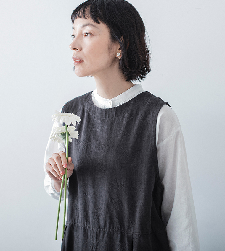 チャコールの2wayオールインワンを白ブラウスに重ね、花を手にした上半身のコーディネート画像