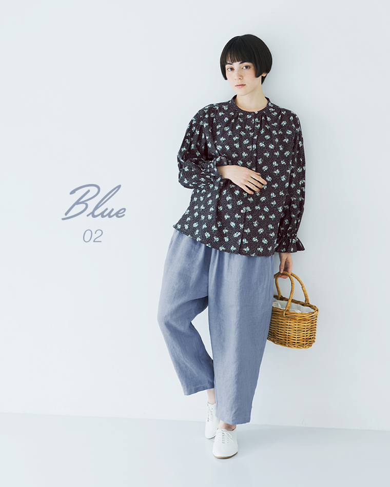 ネイビーの花柄トップスにブルーのパンツを合わせ、かごバッグを持って立つ全身コーディネート画像