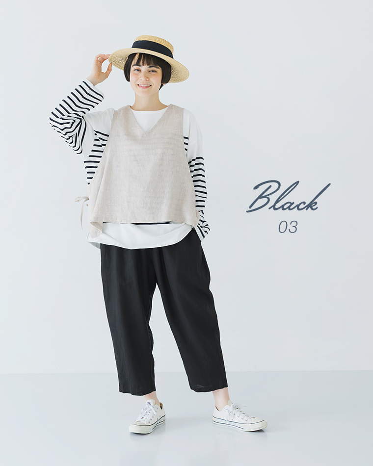 ボーダーの長袖トップスにベージュのベストを重ね、ブラックのパンツと白いスニーカーを合わせ、つば広ハットに手を添えて立つ全身コーディネート画像