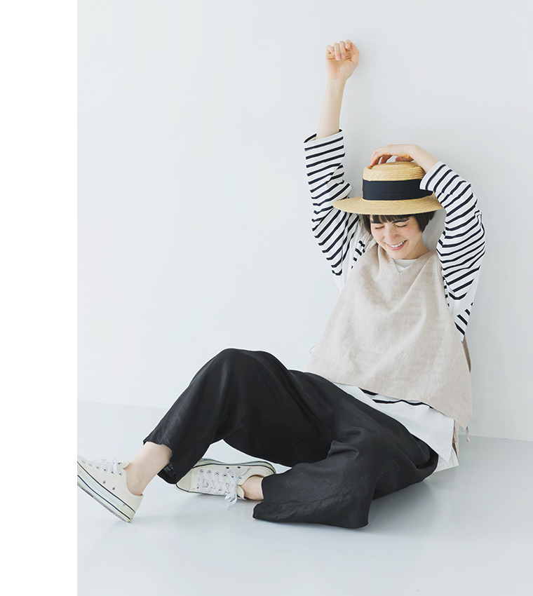 ボーダーの長袖トップスにベージュのベストを重ね、ブラックのパンツと白いスニーカーを合わせ、つば広ハットをかぶって床に座る全身コーディネート画像