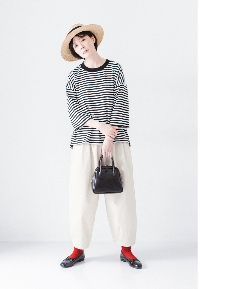 ストローハットをかぶり、ボーダーの長袖トップスにオフホワイトのパンツ、赤い靴下と黒のバレエシューズを合わせ、黒のバッグを持った正面コーディネート