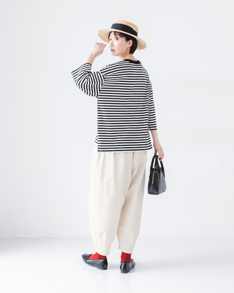 ストローハットをかぶり、ボーダーの長袖トップスにオフホワイトのパンツ、赤い靴下と黒のバレエシューズを合わせ、黒のバッグを持って振り返るポーズのコーディネート