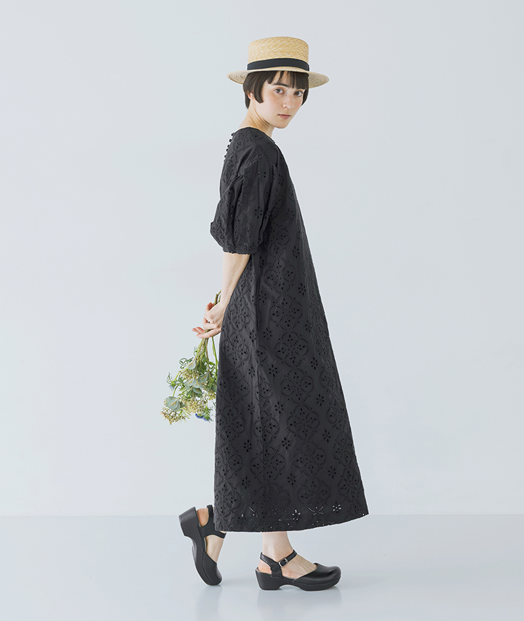 麦わら帽子をかぶったモデルがブラックのカットワークレースワンピースを着て花束を後ろ手に持ち横向きに立っている