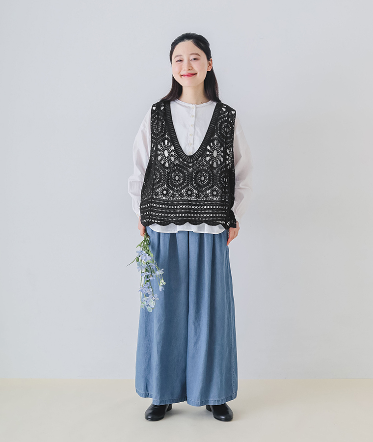 モデルがウエストタックデニムパンツを着てベストとブラウスを合わせ花を持ち正面を向いて立っている