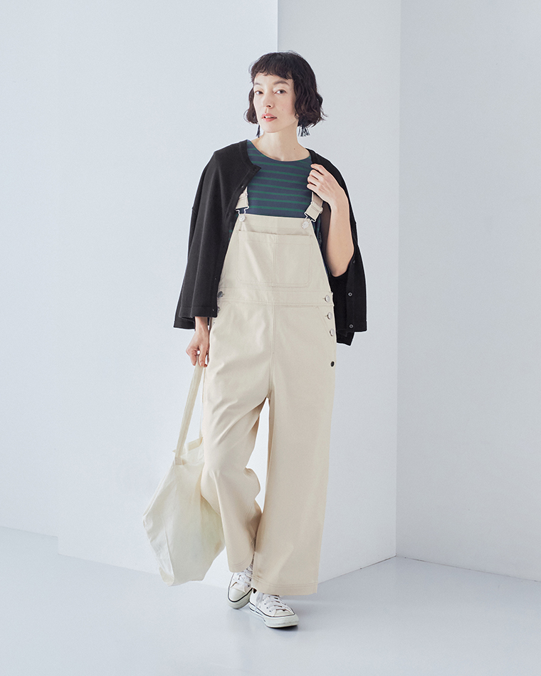 ネイビー×グリーンのボーダー柄トップスにライトベージュのオーバーオールを合わせ、黒の羽織りとオフホワイトのトートバッグを持った全身コーディネート画像