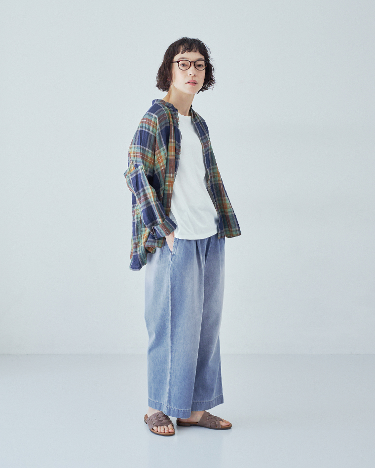 オフホワイトのタンクトップにマルチカラーのチェック柄シャツを羽織り、ライトブルーのワイドパンツとブラウンのサンダルを合わせて座っているコーディネート画像