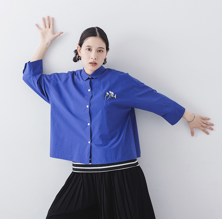 ブルーのシャツに黒のプリーツスカートを合わせた人物が腕を広げて立つ着用コーディネート画像