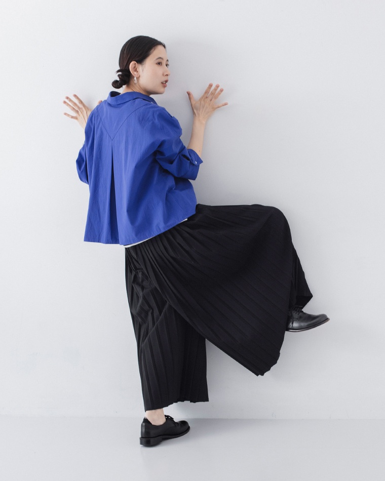 ブルーのシャツに黒のボトムスを合わせた人物が壁に手をつき片脚を上げてポーズを取る着用コーディネート画像