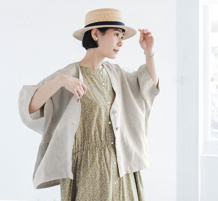 ナチュラルのリネンジャケットを花柄ワンピースに重ね、ストローハットを手で整える着用コーディネート画像