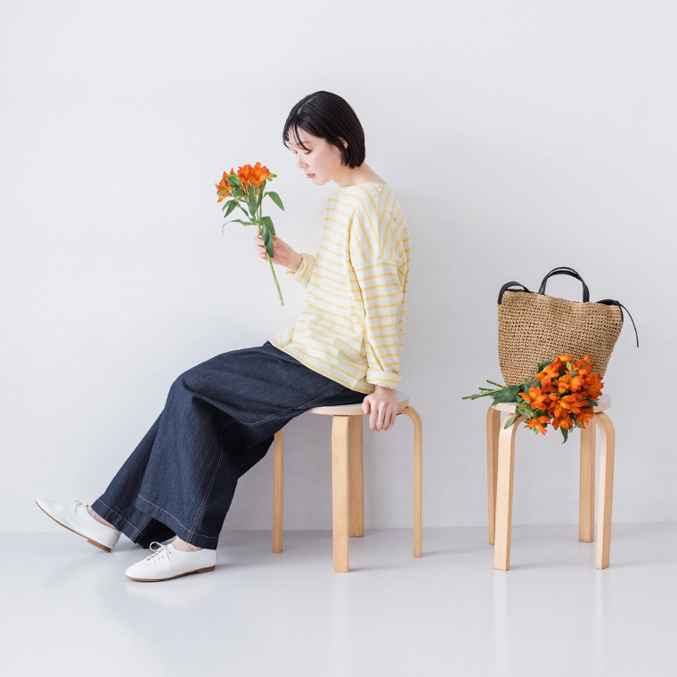 イエロー×ホワイトのボーダートップスにインディゴのワイドデニムパンツと白シューズを合わせ、花を手に持ってスツールに座る着用コーディネート画像