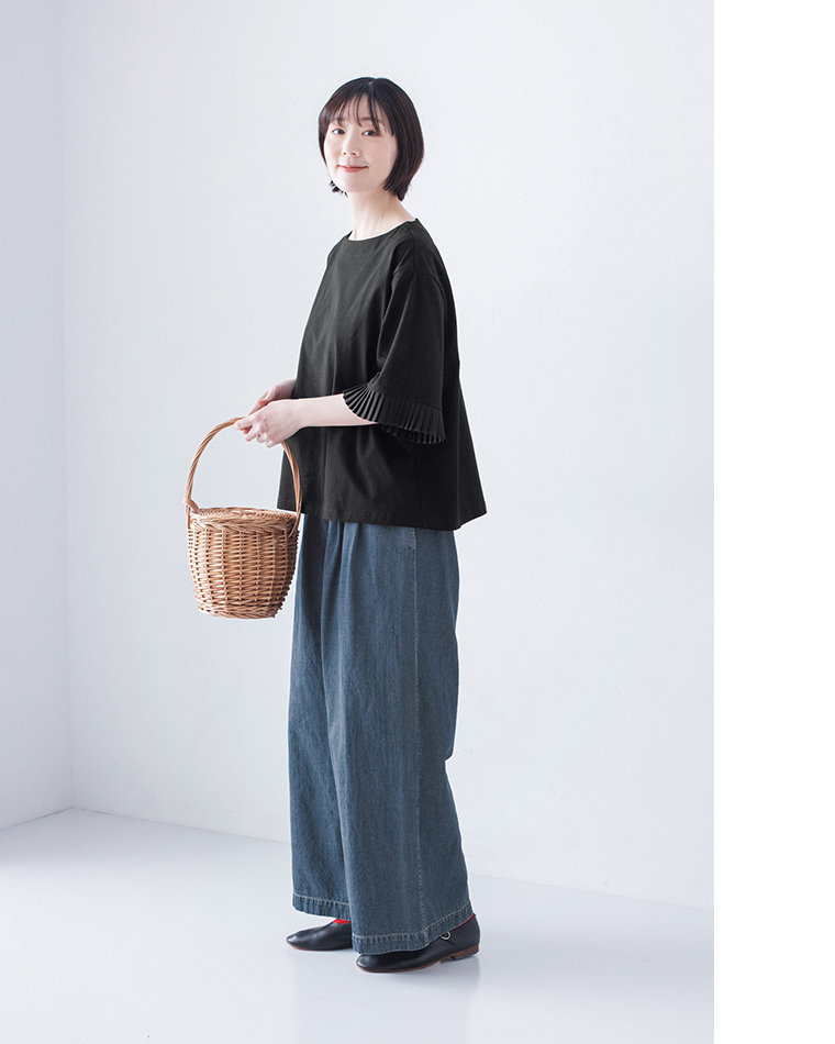 黒のプリーツ袖トップスにインディゴのワイドデニムパンツと黒シューズを合わせ、丸いかごバッグを手に持つ着用コーディネート画像