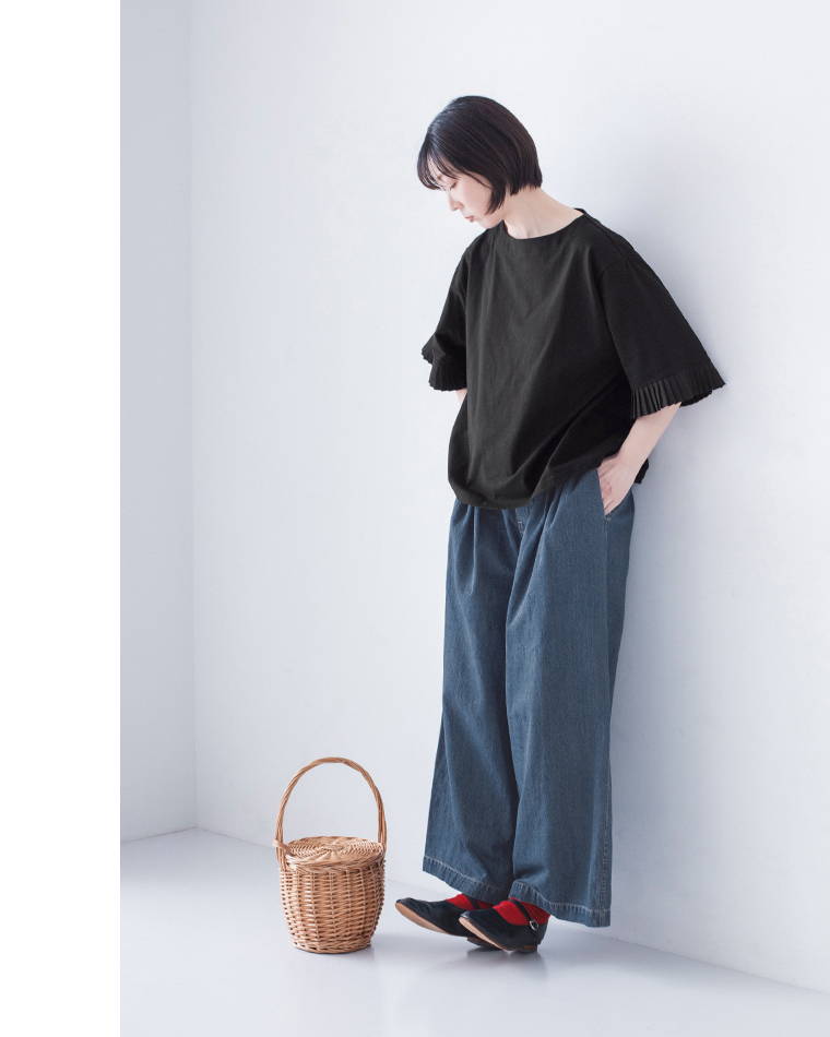 黒のプリーツ袖トップスにインディゴのワイドデニムパンツと黒シューズを合わせ、足元に丸いかごバッグを置いた着用コーディネート画像