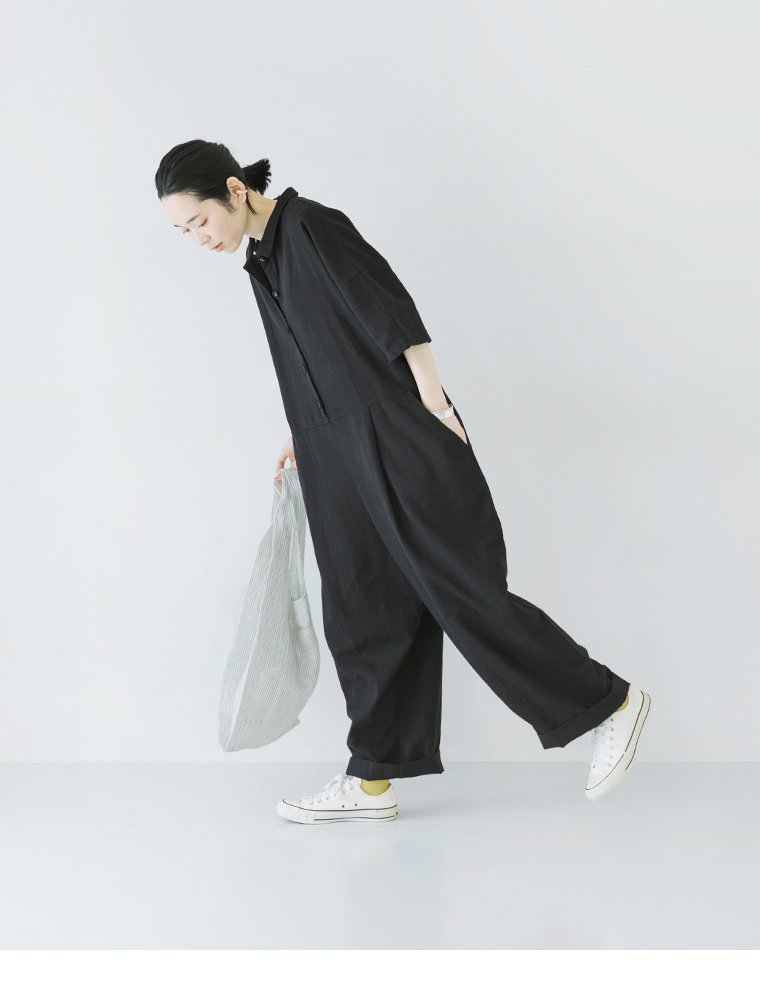 ブラックのオールインワンを着用した人物がバッグを持って前傾姿勢で立つ全身コーディネート写真