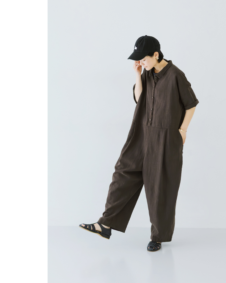 ブラウンのオールインワンを着用した人物が片足を軽く上げて立つ全身コーディネート写真