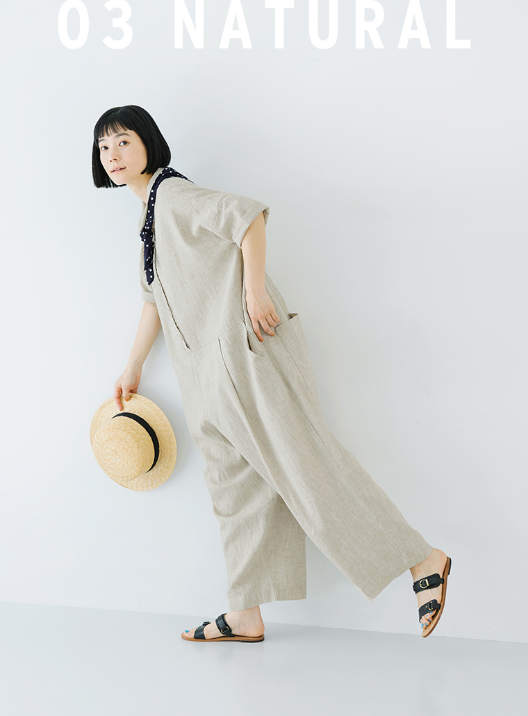 ナチュラルのオールインワンを着用した人物が帽子を手に片足を軽く上げて立つ全身コーディネート写真（03 NATURAL）