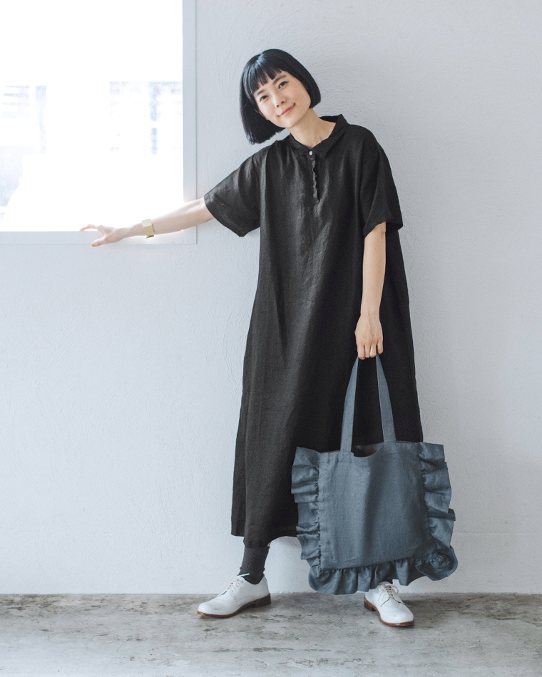 08Mabの リネンフリルトートバッグ を手にした女性のコーディネート画像。黒のリネンワンピースに白いレースアップシューズを合わせ、グレーのフリルトートを持って立つ姿。