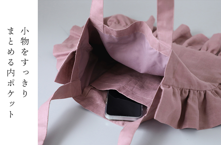 08Mabの リネンフリルトートバッグ（ダスティーピンク）の内ポケットにスマートフォンを入れた様子。フリルの縁取りが見える開口部のアップで、収納の便利さを伝える構図。画像内テキスト「小物をすっきりまとめる内ポケット」。