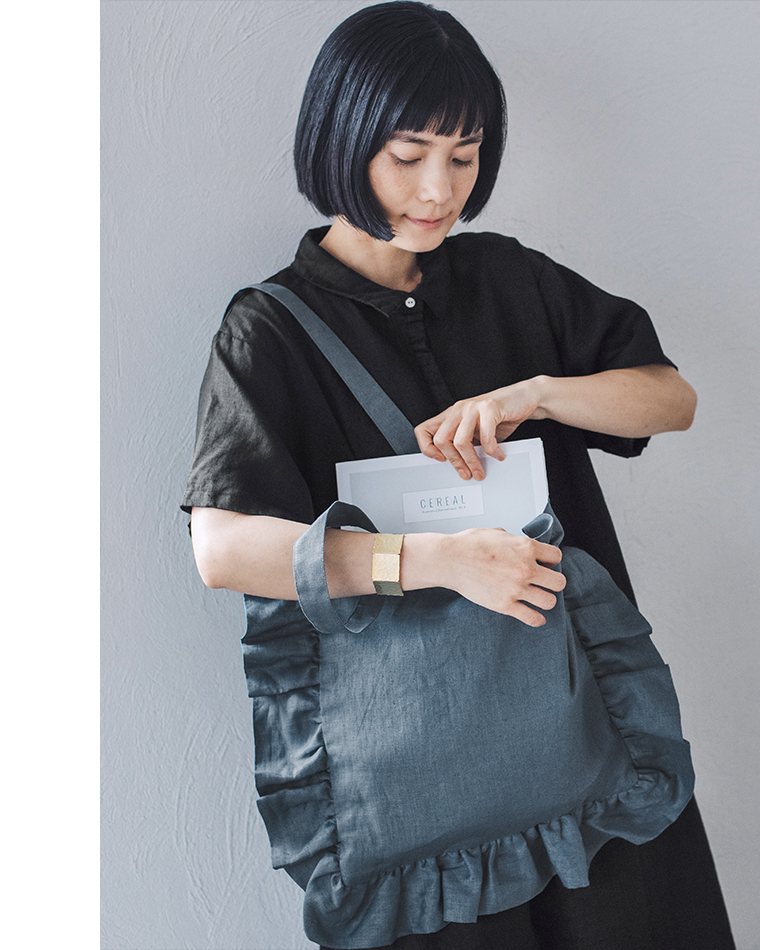 08Mabの リネンフリルトートバッグ（ダルブルー）を肩に掛けた女性のコーディネート画像。黒のリネンワンピースに合わせ、雑誌「CEREAL」をバッグに入れる様子。ナチュラルで落ち着いたトーンが印象的なスタイル。