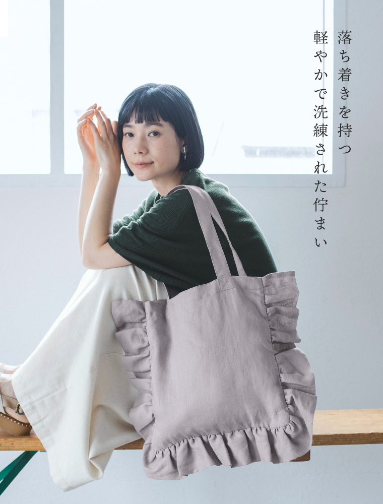 08Mabの リネンフリルトートバッグ（スチールグレー）を肩に掛けた女性のコーディネート画像。グリーンのトップスに白のワイドパンツを合わせ、ナチュラルな雰囲気で座る姿。画像内テキスト「落ち着きを持つ 軽やかで洗練された佇まい」。