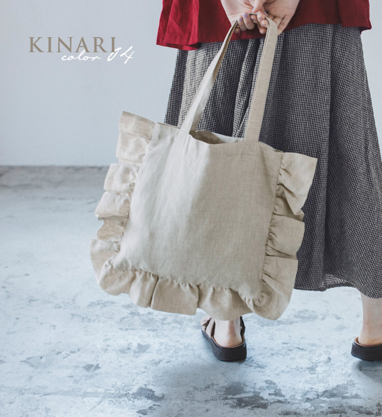 08Mabの リネンフリルトートバッグ（キナリ）を手にした女性のコーディネート画像。赤のトップスにチェック柄のロングスカートを合わせ、ナチュラルなトーンでまとめたスタイル。画像内テキスト「KINARI color 04」。