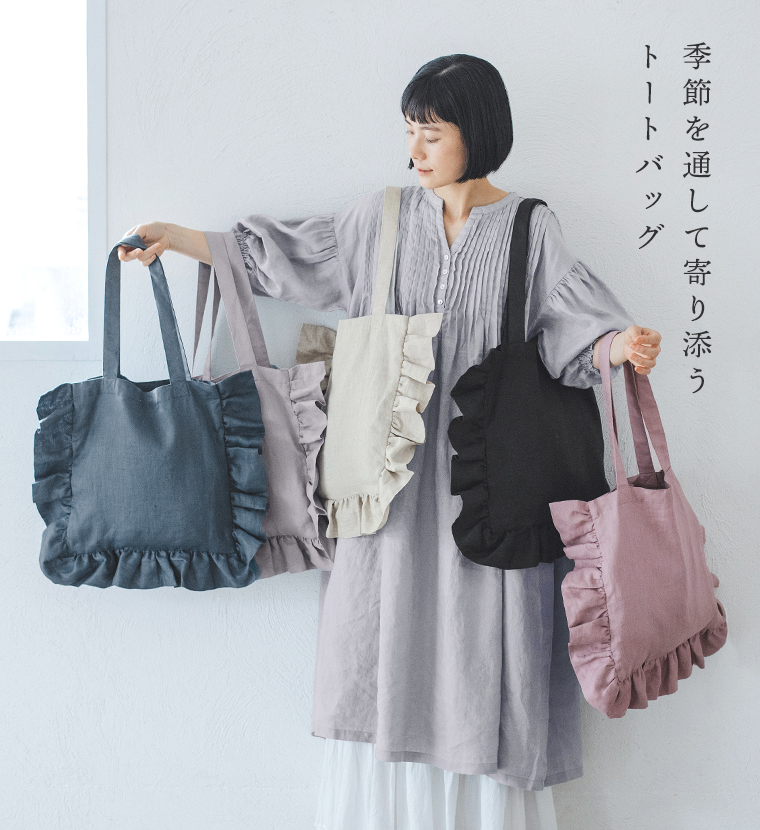 08Mabの リネンフリルトートバッグ（ブラック）を手にした女性のコーディネート画像。ライトグレーのピンタックワンピースに白のペチスカートを重ね、黒のサンダルを合わせたナチュラルスタイル。画像内テキスト「季節を通して寄り添うトートバッグ」。