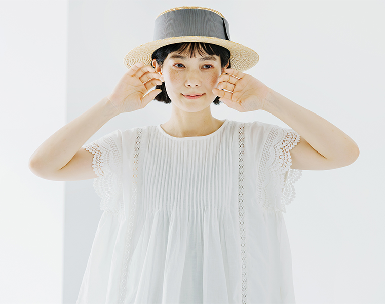 Lintu Lauluレースピンタックブラウス（ホワイト）の着用イメージ。繊細なピンタックとスカラップレース袖が特徴のブラウスを、麦わら帽子と合わせた爽やかなスタイリング。軽やかなコットン素材がふんわりと広がり、夏らしい清涼感を表現。
