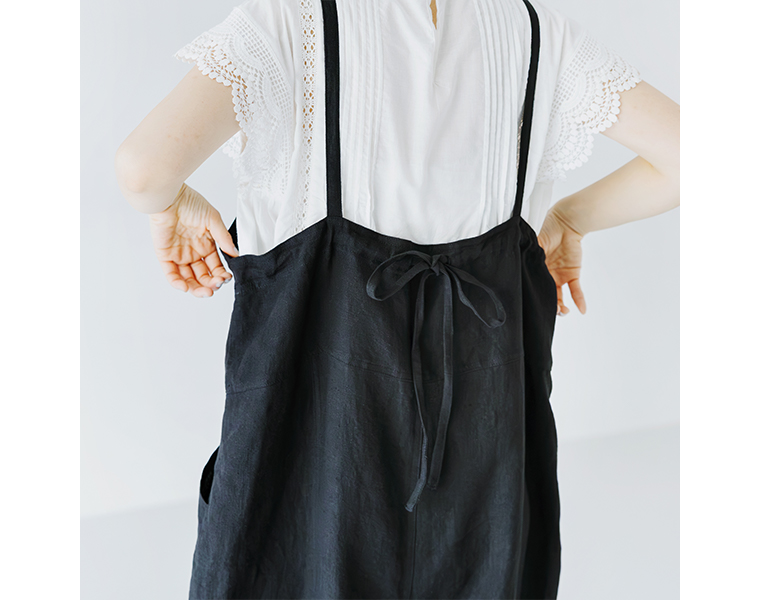&yarn100％リネンバレルサロペット（ブラック）の後ろ姿ディテール画像。繊細なレースブラウスに合わせ、背中のリボン結びと細ストラップのデザインを写したカット。リネン素材の柔らかな質感と上品な印象を表現。