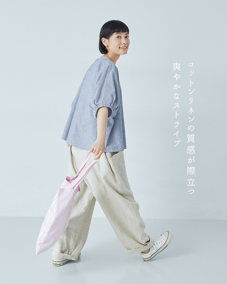 Bliss bunchのちょうちんスリーブブラウス（ブルーストライプ）を着用したモデルの横向きカット。白のサロペットと白のスニーカーを合わせ、ピンクのロゴ入りトートバッグを持って歩く姿。右側に「コットンリネンの質感が際立つ　爽やかなストライプ」というテキスト入りで、軽やかな素材感を表現したスタイリング。