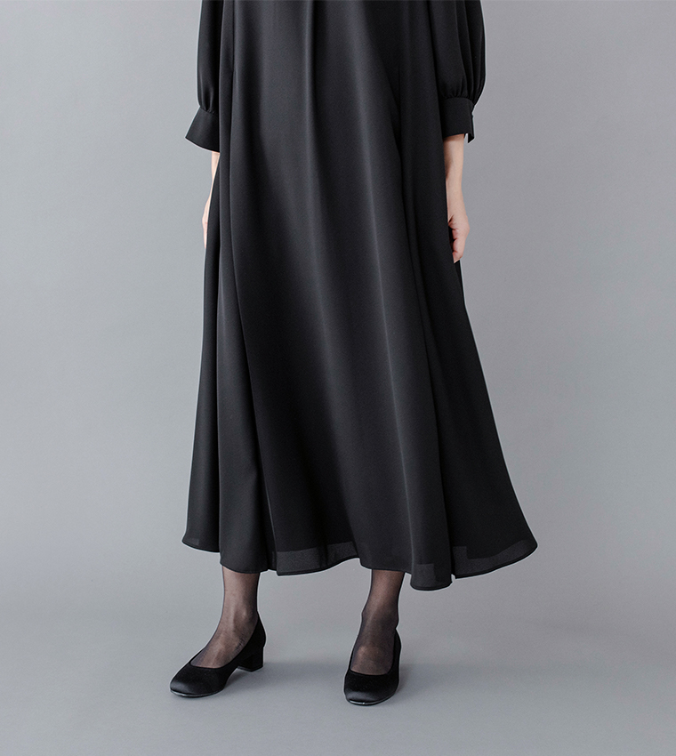 Luuna miu【慶弔両用】大切な日のボタンフレアワンピースの裾ディテールイメージ。黒のロングワンピースを着たモデルの立ち姿で、裾がふんわりと広がる上品なシルエットを強調。軽やかな生地の流れと足元の黒パンプスがフォーマルな印象を引き立てる商品画像。