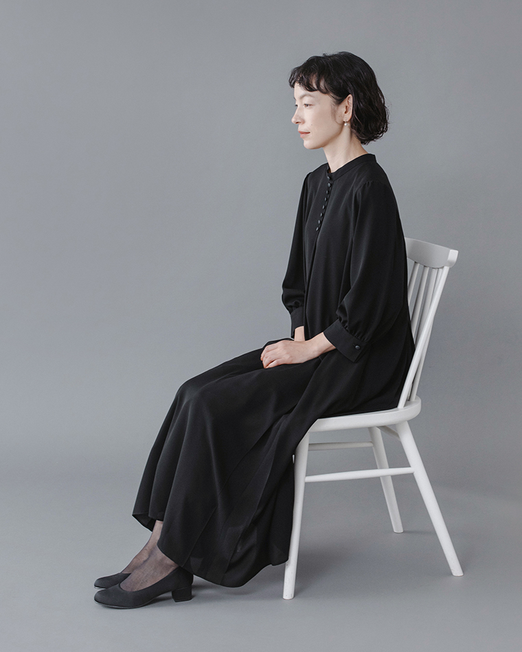 Luuna miu【慶弔両用】大切な日のボタンフレアワンピース着用イメージ。黒のロングワンピースを着たモデルが白い椅子に座り、横向きで静かに手を膝に置いている姿を写した写真。シンプルなデザインと柔らかな生地の流れが上品な印象を与えるフォーマルスタイルの商品画像。