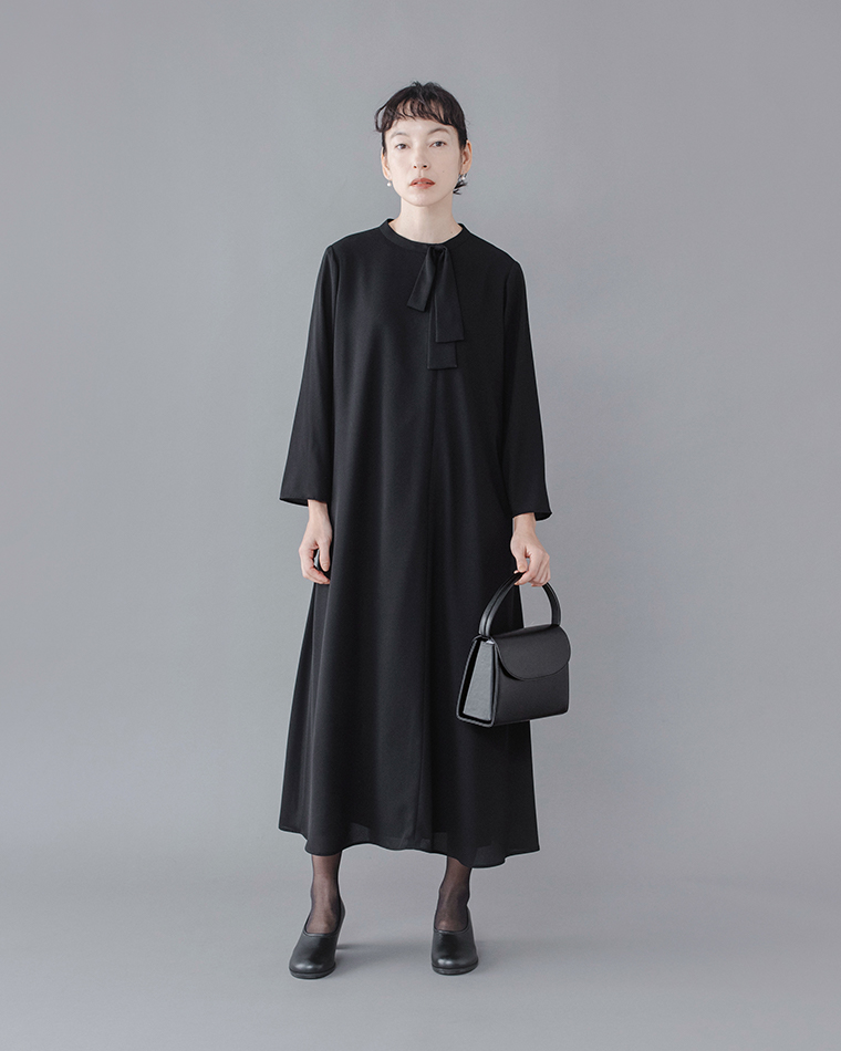 Luuna miu【慶弔両用】大切な日のボウタイAラインワンピース着用イメージ。黒のロングワンピースを着たモデルがグレー背景の前に立ち、首元のボウタイが上品に揺れる。黒のパンプスとハンドバッグを合わせたフォーマルスタイルで、シンプルながらも優雅なシルエットが際立つ商品画像。