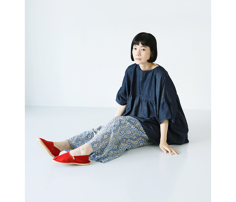 Sorte cuffぜいたくギャザーワイドブラウス（インディゴ）を着用した女性の座り姿コーディネート。深みのあるインディゴカラーが印象的で、ゆったりとしたギャザーシルエットが軽やかな印象を与える。柄パンツと赤いシューズを合わせたリラックス感のあるスタイリングの商品画像。