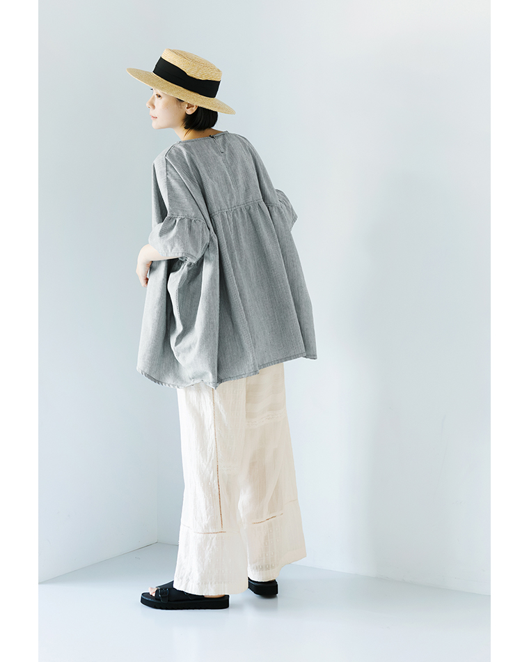 Sorte cuffぜいたくギャザーワイドブラウス（ヒッコリー）を着用した女性の後ろ姿コーディネート。ほどよくハリのあるヒッコリー素材が爽やかな印象を与え、ふんわりと広がるギャザーが軽やか。麦わら帽子と白のパンツを合わせたナチュラルで涼しげなスタイリングの商品画像。