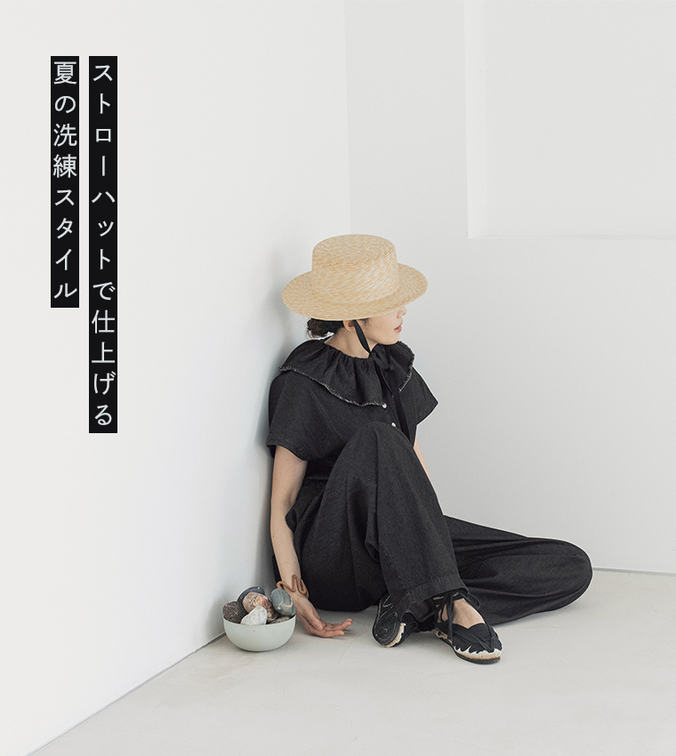 &yarn 滝口和代さんコラボ　or frill all in oneを着用したイメージ。黒のフリル付きオールインワンにストローハットを合わせた、夏らしく洗練されたスタイル。白い空間で軽やかに座る姿がナチュラルな雰囲気を引き立てる商品画像。