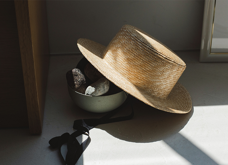 田中帽子店 滝口和代さん×田中帽子店コラボ quiet straw hat のディテールイメージ。自然光の差し込む室内で、黒いリボン付きの麦わら帽子が石を入れた器に軽く掛けられている様子。素材の質感と静かな空気感が伝わる構図。