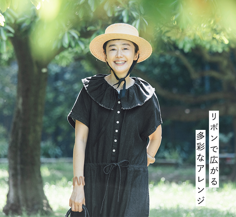 田中帽子店 滝口和代さん×田中帽子店コラボ quiet straw hat を着用した人物の屋外イメージ。黒いリボンを結んだ麦わら帽子と黒のワンピースを合わせ、緑の木々の下で微笑む姿。自然光に包まれた穏やかな雰囲気が伝わるスタイリングカット。